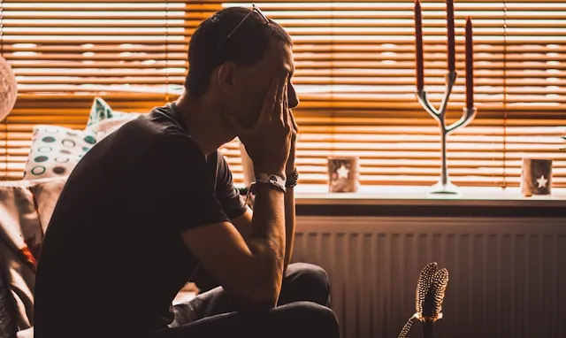 A man sits by a sunlit window with blinds, head in his hands, looking down.